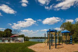 Children's play park with woodchips, picnic table, fenced in pool, and lake in background